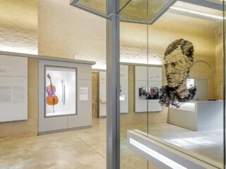 Museumsraum mit verschiedenen Vitrinen und Objekten. Im Vordergrund ein Spiegel mit dem verpixelten Kopf von Claus Schenk Graf von Stauffenberg.