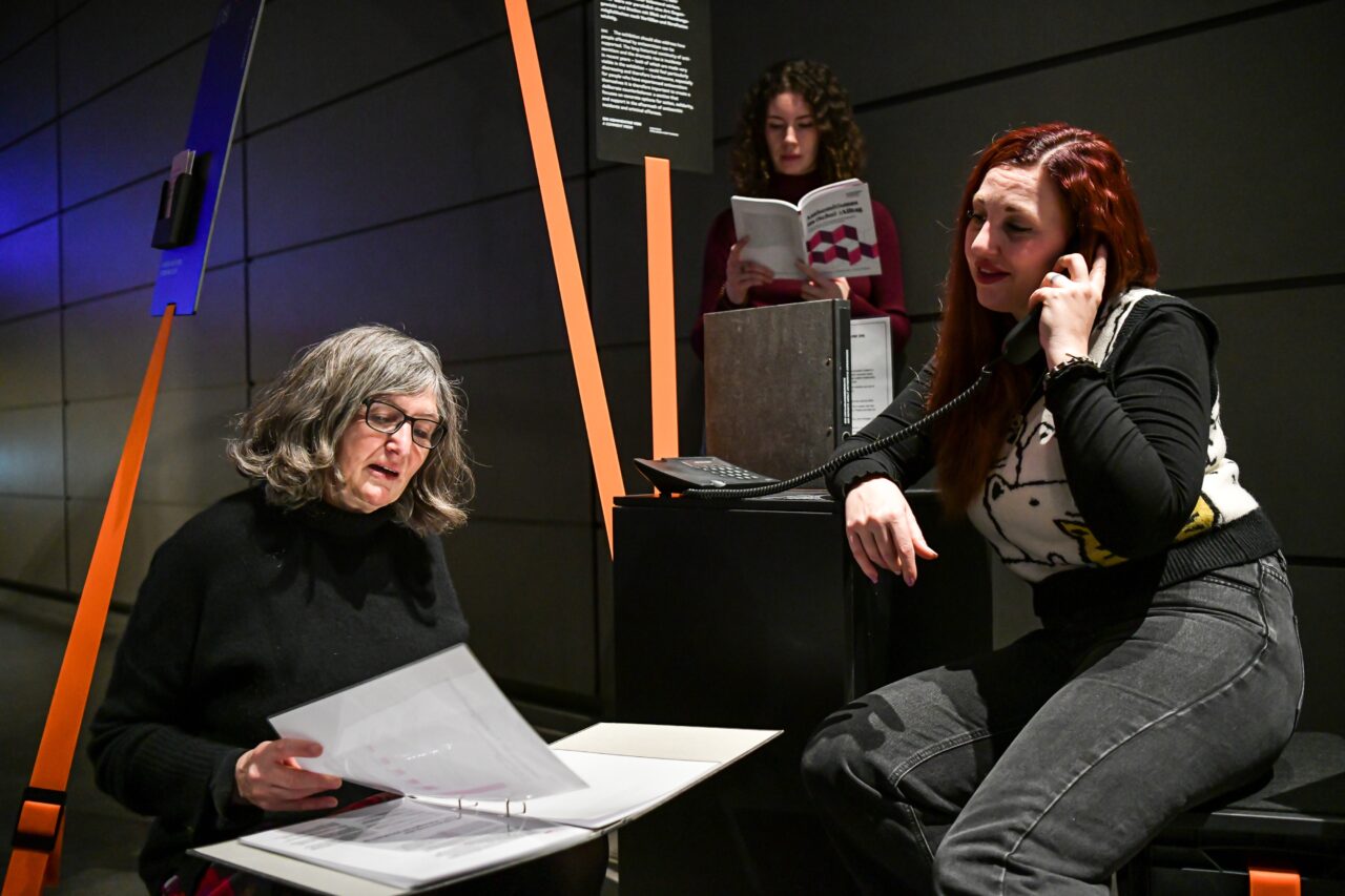 Zwei Frauen lesen Informationsmaterialien, eine hört am Telefon zu.